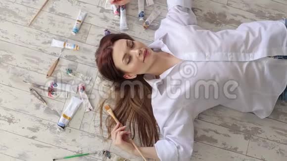 女孩艺术家躺在地板上在工作室的许多油漆管中视频的预览图