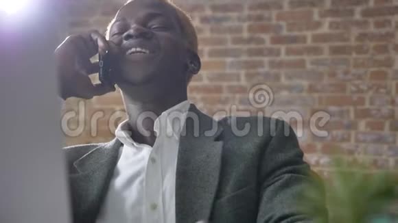 年轻的非洲快乐金发商人打电话坐在现代办公室笑着笑着视频的预览图