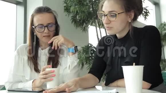 两个戴着眼镜的年轻商务女孩坐在咖啡馆的桌子旁喝着咖啡讨论着商业想法视频的预览图