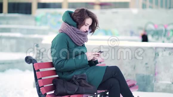 一个快乐的女人在冬天的城市里坐在长椅上对着满是涂鸦的石块发短信视频的预览图