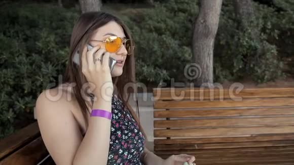 一个年轻的女孩坐在公园里的长凳上喝水打电话夏季和自然视频的预览图