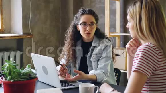 年轻的女企业家正和她的伴侣在现代办公室交谈同时坐在办公桌前女人在计划新的视频的预览图
