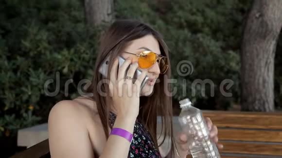 一个年轻的女孩坐在公园里的长凳上喝水打电话夏季和自然视频的预览图