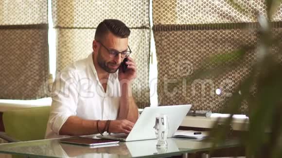成熟的男人穿着白色衬衫留着胡子坐在笔记本电脑前一边打电话开心地笑挂起来视频的预览图