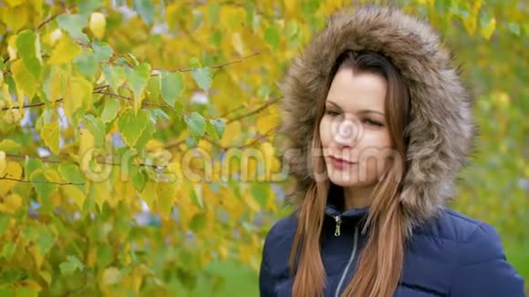 女孩在秋天的城市公园散步穿夹克衫的年轻女子在秋树背景上戴着毛皮帽女孩走在里面视频的预览图