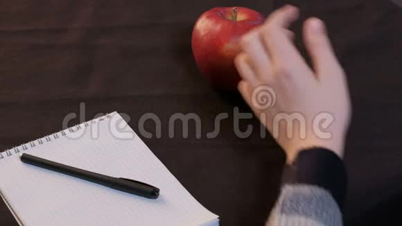特写镜头一个红苹果躺在记事本和黑色布上的钢笔旁边一个年轻人的手从盘子里拿起一个成熟的水果视频的预览图
