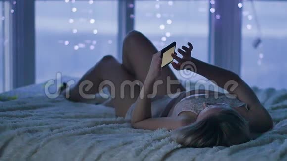迷人的金发女郎在她的智能手机上写信息躺在床上卧室里拿着手机的年轻女子视频的预览图