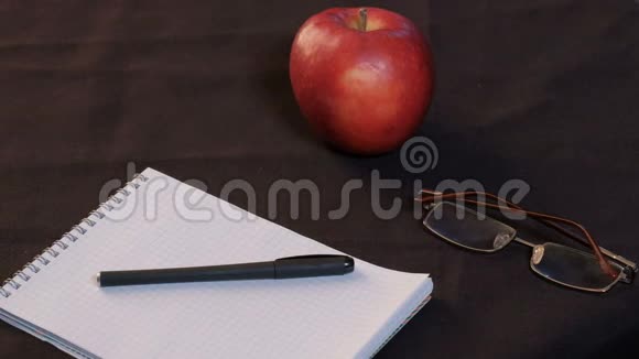 特写镜头一个人从桌子上举起眼镜一个红苹果躺在记事本和黑色布上的钢笔旁边视频的预览图