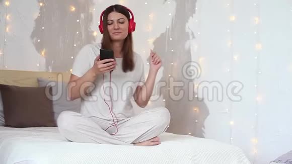 美丽的女孩听音乐耳机红色坐在床上放松使用手机高清高清视频的预览图
