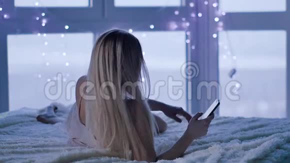 迷人的金发女郎在她的智能手机上写信息躺在床上卧室里拿着手机的年轻女子视频的预览图