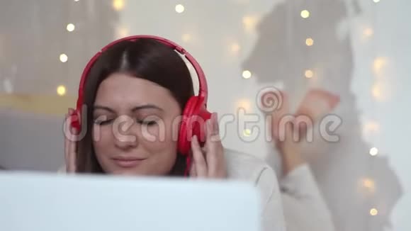美丽的快乐女孩躺在床上用白色的移动电脑听耳机上的音乐高清特写视频的预览图