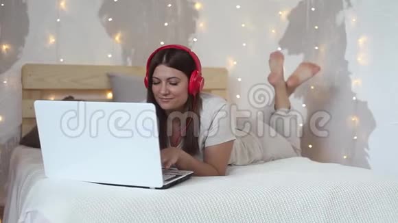 美丽的快乐女孩躺在一张大床上用白色的电脑移动电脑和耳机听音乐视频的预览图