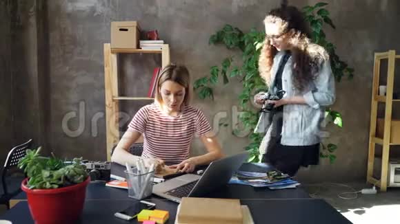 年轻的设计师正坐在桌旁的笔记本上画照片而女摄影师正带着相机来看她视频的预览图