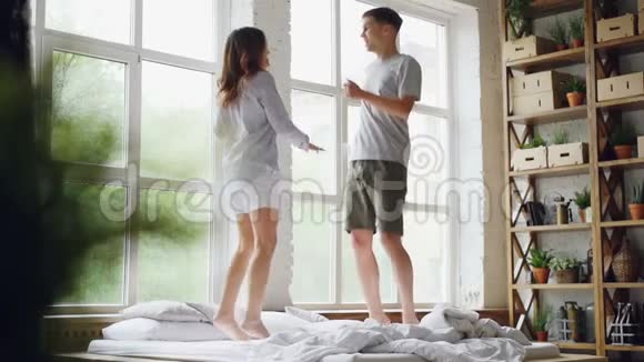 新婚夫妇在有大窗户的小房间里的双人床上跳跃和跳舞的动作很慢快乐的视频的预览图