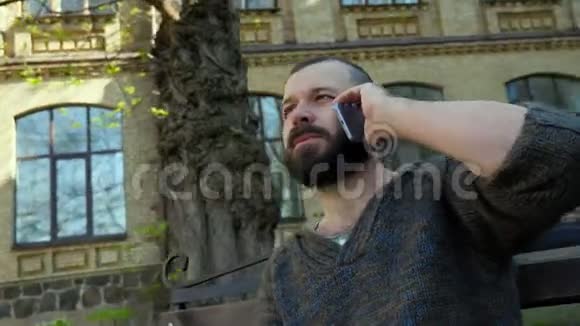 这名留胡子的男子坐在城市公园的长凳上在旅途中打电话视频的预览图