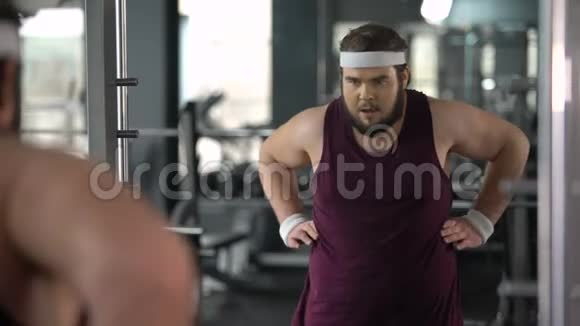 有趣的胖子看着镜子反射健身房摆姿势假装肌肉发达视频的预览图