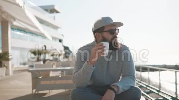 快乐的大胡子男人在海边咖啡馆的露台上用纸杯喝咖啡视频的预览图