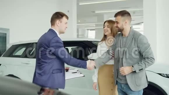 开朗的汽车经销商销售员正和年轻的买主握手帅哥然后把钥匙交给他快乐的妻子视频的预览图