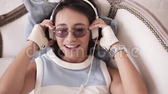 可爱的开朗的女孩戴着耳机和眼镜一边躺在沙发上一边微笑着和音乐一起跳舞视频的预览图