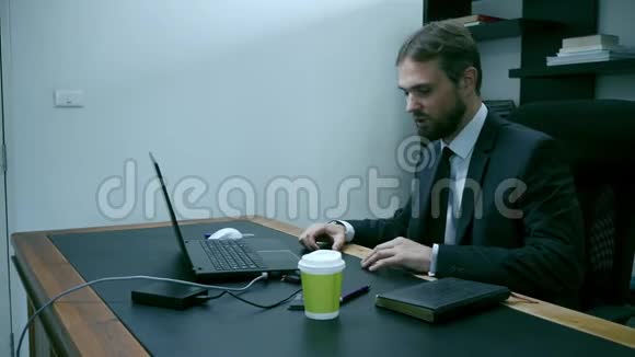 坐在笔记本电脑后面的办公室里一个穿着衣服的商人男工看着手机很生气很慢视频的预览图