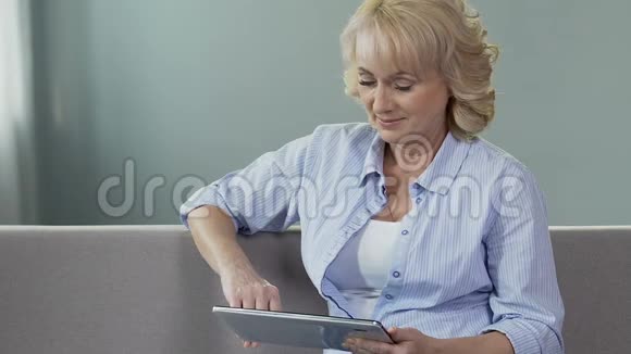 沙发上的老年妇女滚动着平板电脑屏幕微笑着看着照片视频的预览图