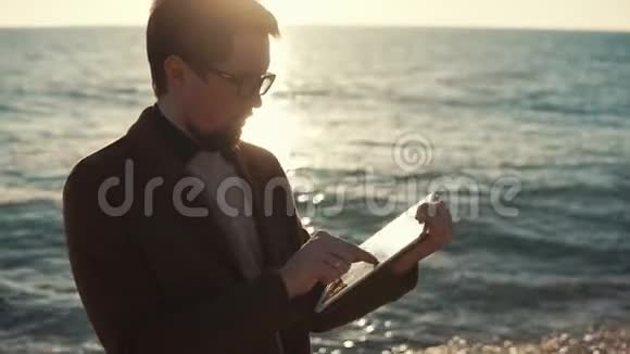 一位在便携式平板电脑上工作的成年人站在海边的商人视频的预览图