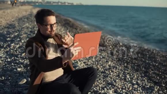 微笑的男人正在鹅卵石海滩上用平板电脑做视频通话视频的预览图