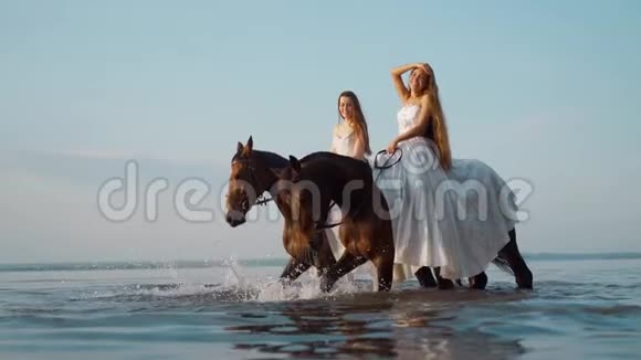 两个穿着白色长裙的漂亮女孩骑马骑马的人在日落时沿着海岸漫步视频的预览图