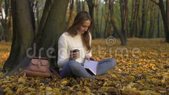 可爱的女孩坐在公园的树下喝着咖啡读着日记视频的预览图