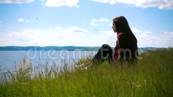 在阳光明媚的日子里坐在山上看着河的体贴的年轻女人视频的预览图