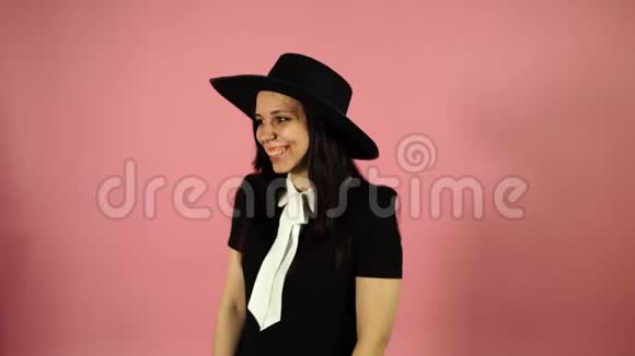 女士在粉红色的背景上微笑穿黑色连衣裙和帽子的女人视频的预览图