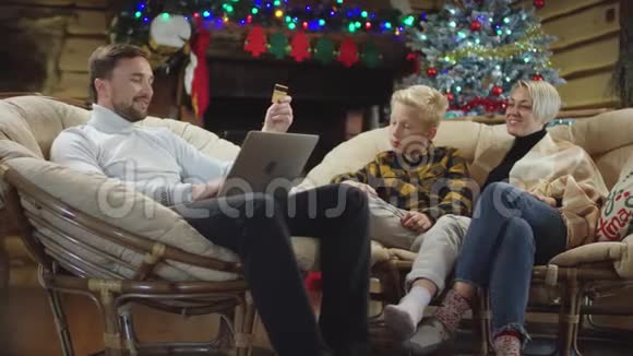 有信用卡的男人在笔记本电脑上购物而小圣诞礼物落在他的妻子和儿子身上视频的预览图