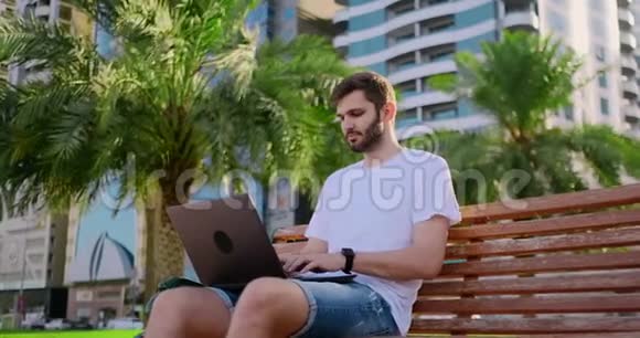 在棕榈树的夏天一个年轻人拿着笔记本电脑坐在键盘上打字视频的预览图