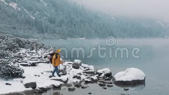 徒步旅行者穿着黄色的冬衣走在靠近山湖的石头上举起双手在山顶上获胜视频的预览图