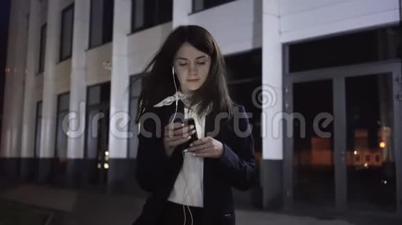 一位年轻的女商人蒙太奇在一个夜城里戴着智能手机和耳机视频的预览图