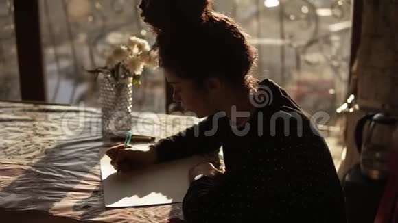 可爱的女艺术家她的卷发挽着发髻坐在艺术工作室全景窗口附近的桌子旁视频的预览图