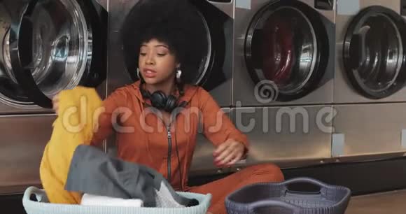 快乐的非洲裔美国年轻女性在洗衣前用篮子整理衣物然后坐在自助服务中洗衣服视频的预览图