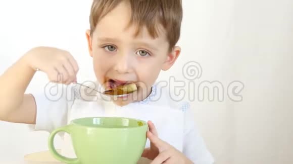孩子高兴地用勺子从盘子里吃汤特写一个男孩坐在白色背景的桌子旁吃饭视频的预览图