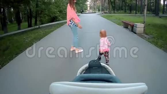 快乐的年轻妈妈和小女孩在夏天公园跑步从一个带着婴儿车的父亲眼中视频的预览图