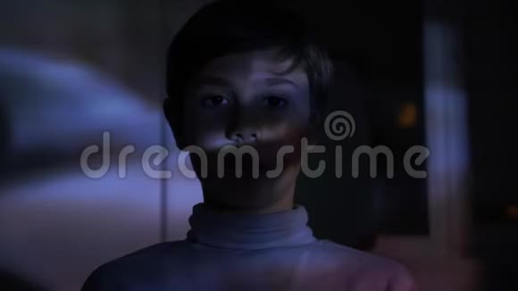 小男孩在黑暗的房间里看恐怖电影或电视或电脑视频他脸上的倒影视频的预览图