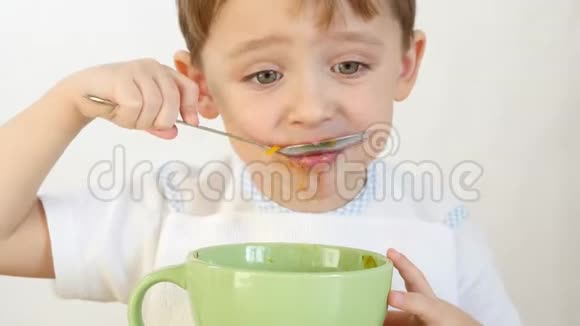 一个快乐的孩子用勺子特写从盘子里吃汤那个男孩坐在一张白色的桌子旁吃饭视频的预览图