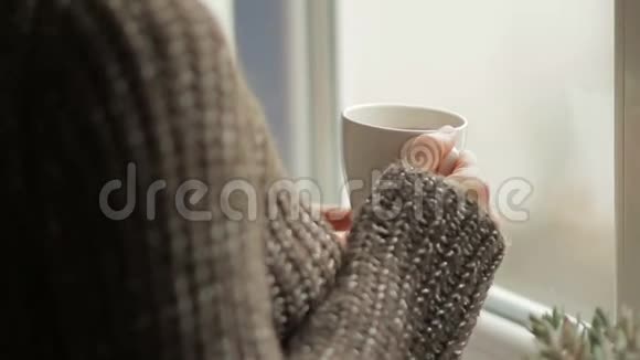 一位穿着暖和毛衣的女士站在靠窗的舒适的房子里喝着热饮视频的预览图