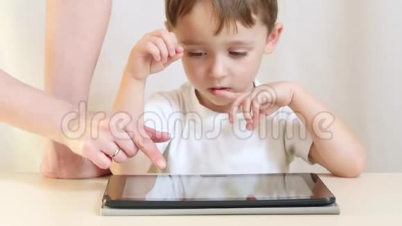 一个快乐的孩子坐在桌子旁在平板电脑上玩耍妈妈手帮孩子在平板上玩孩子视频的预览图