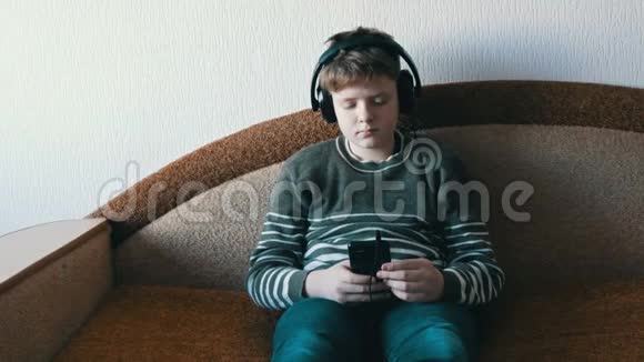 头上戴着大耳机的少年正坐在棕色沙发上在智能手机上听音乐视频的预览图
