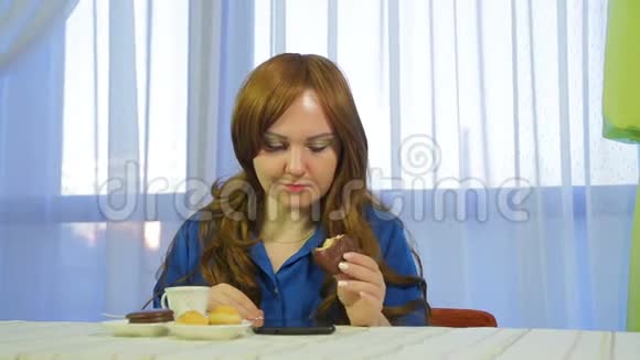 年轻的棕色头发的女人在一张桌子上的咖啡馆里喝着咖啡和甜点读着平板电脑上的新闻视频的预览图
