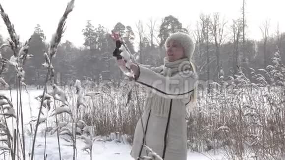这位穿着白色夹克的年轻女士在智能手机上拍摄了冬季森林湖海岸的全景它是视频的预览图