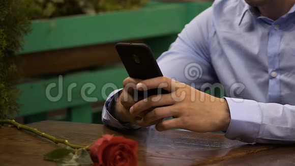 男人用手机坐在桌边红玫瑰躺在前面等待约会视频的预览图
