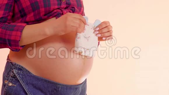 孕妇手里拿着新生的帽子戴在她赤裸的大肚子上怀着可爱婴儿衣服的孕妇视频的预览图