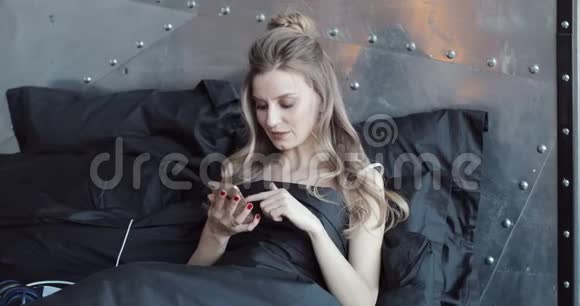漂亮的金发女郎在黑色毯子下坐在床上检查她的手机视频的预览图