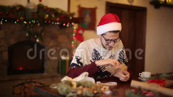 戴着圣诞帽的快乐男人在壁炉旁打开圣诞礼物盒里面有闪烁的花环微笑的家伙收到了视频的预览图